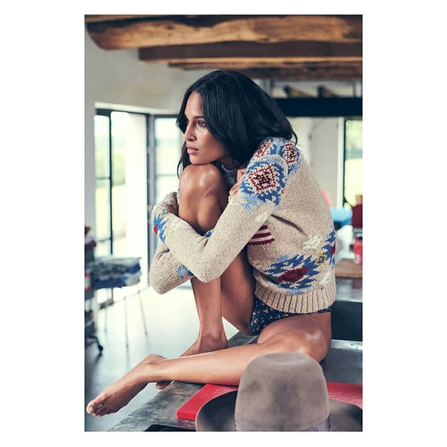 Adult female in a beige patterned sweater sitting on counter with knees up, looking off into the distance in an indoor environment.