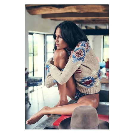 Adult female in a beige patterned sweater sitting on counter with knees up, looking off into the distance in an indoor environment.