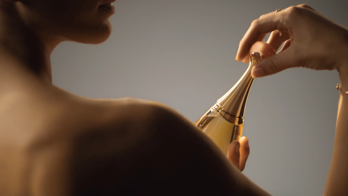 Woman's bare back and shoulders, holding a golden perfume bottle in her hand, against a neutral grey backdrop in a beauty advertising style.
