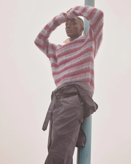 A young black man in a striped sweater leans against a blue post outdoors under soft light, creating a calm, fashion-focused portrait.