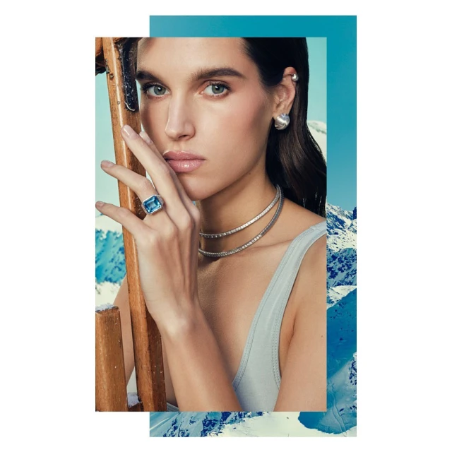 Close-up of a brunette woman wearing jewelry, including a ring and necklace, with a snowy mountain landscape and blue sky in the background.