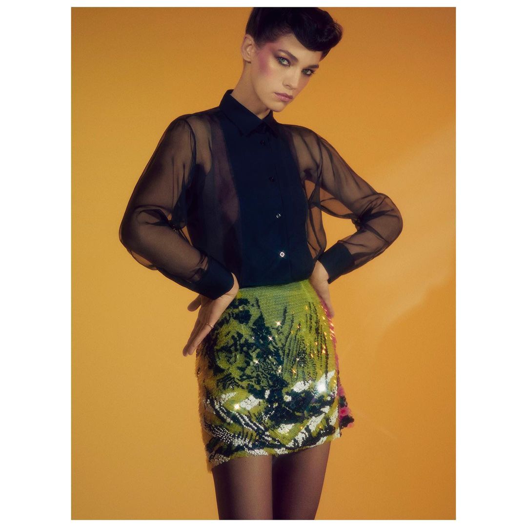 Fashionable woman in black sheer blouse and bright sequined mini skirt poses against a yellow backdrop in a studio portrait.