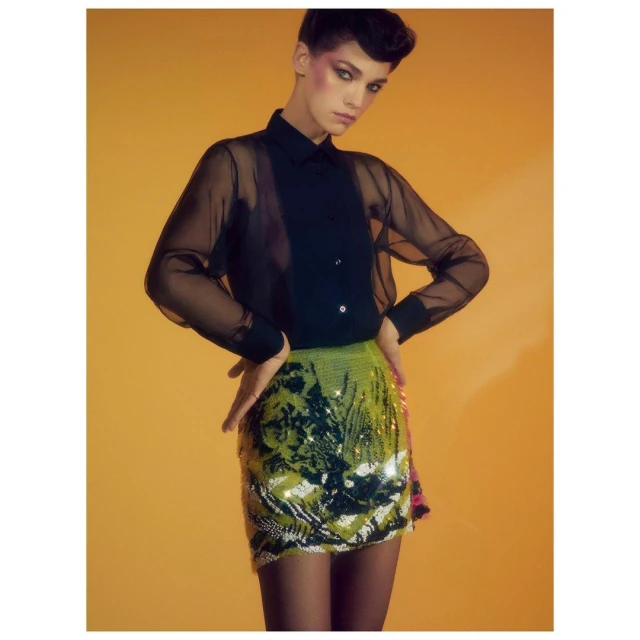 Fashionable woman in black sheer blouse and bright sequined mini skirt poses against a yellow backdrop in a studio portrait.