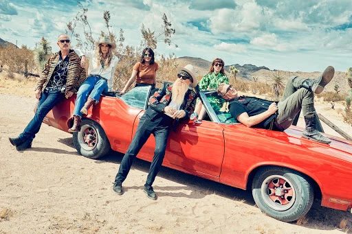 Group of six people posing with a red convertible car in a desert landscape with mountains under a partly cloudy sky, daytime.
