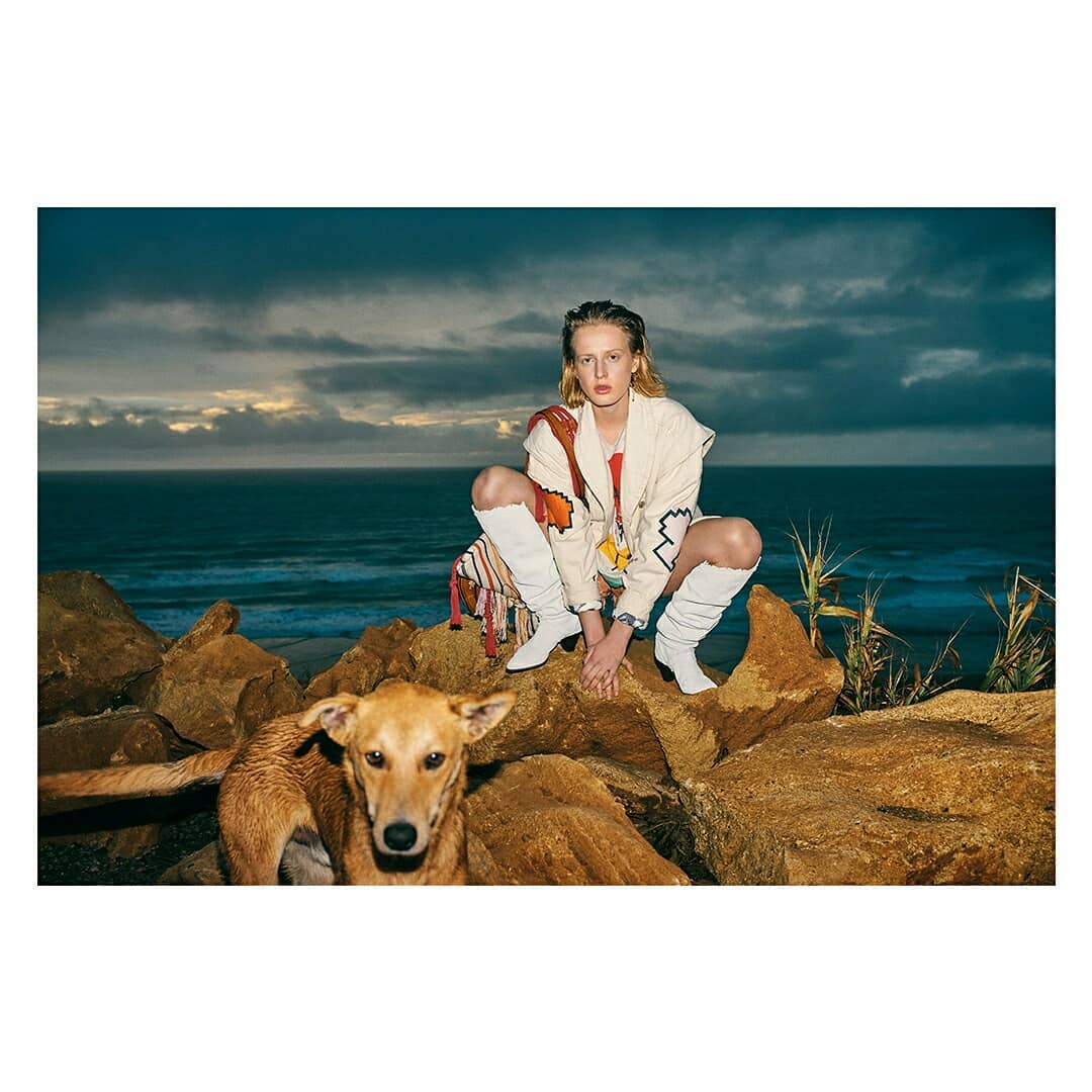 A blonde woman wearing a jacket and white boots is crouching on rocks next to a brown dog with a blue ocean and sky background.