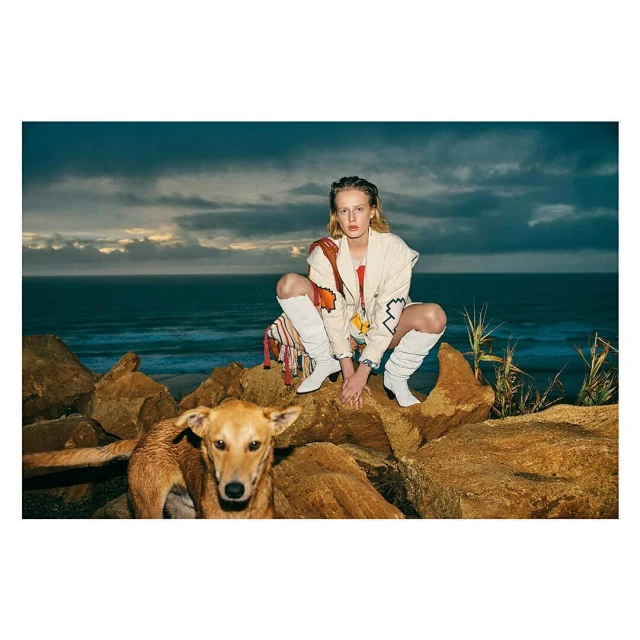 A blonde woman wearing a jacket and white boots is crouching on rocks next to a brown dog with a blue ocean and sky background.