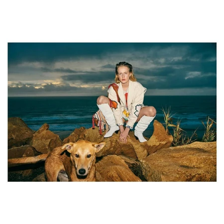A blonde woman wearing a jacket and white boots is crouching on rocks next to a brown dog with a blue ocean and sky background.