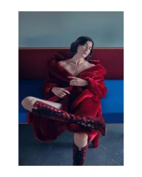 Woman in a plush red faux fur coat and knee-high boots seated against a blue and maroon bench interior.