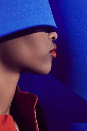 Close-up profile of a Black woman wearing a blue visor and red jacket against a vibrant blue textured background.