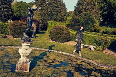 Model walking beside an ornamental garden pond featuring a classical statue and manicured hedges in sunlight.