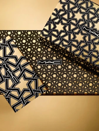 Geometric pattern of tessellated black stars on gold cosmetic product packaging featuring the MAC logo in the center of the image.