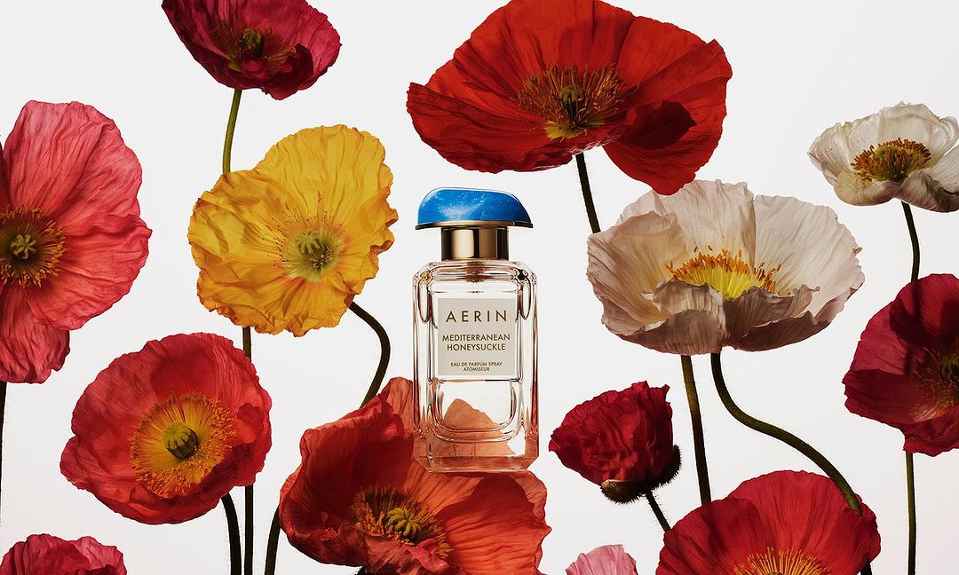 AERIN Mediterranean Honeysuckle perfume bottle surrounded by red, yellow, and white poppies against a bright white background.