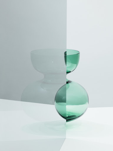 Minimalist still life with a green glass bowl stacked on a green glass sphere, set against a two-tone gray and white backdrop.
