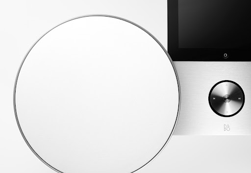 Close-up of a modern audio device with a round speaker and sleek interface on a white surface, minimalist design, and B&O logo.