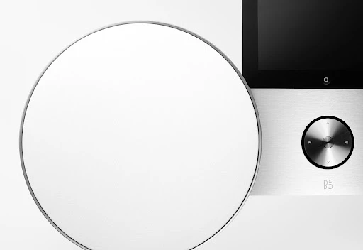 Close-up of a modern audio device with a round speaker and sleek interface on a white surface, minimalist design, and B&O logo.
