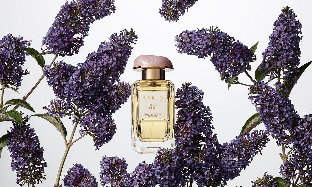 Aerin Lilac Path perfume bottle with a pink cap surrounded by lilac blossoms against a white background in a beauty still life.