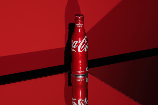 Studio shot of a red Coca-Cola bottle reflected on a glossy surface against a matching red background with contrasting shadows.