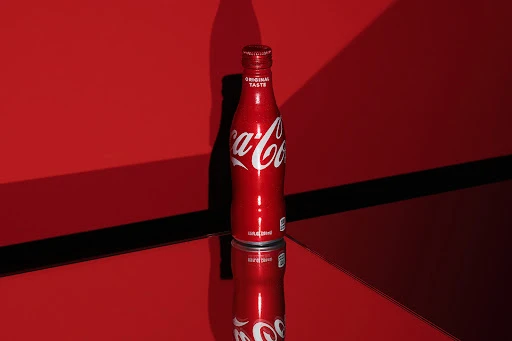 Studio shot of a red Coca-Cola bottle reflected on a glossy surface against a matching red background with contrasting shadows.