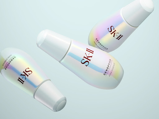 Three SK-II GenOptics Aura Essence bottles in a studio setting, showcasing skincare product and cosmetic brand promotion on a light blue background.