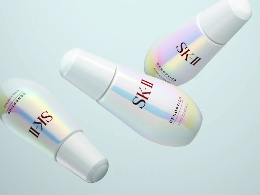 Three SK-II GenOptics Aura Essence bottles in a studio setting, showcasing skincare product and cosmetic brand promotion on a light blue background.