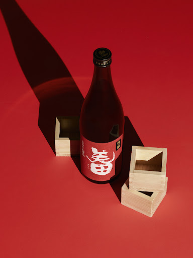 Bottle of sake with wooden square cups on red background. Shadows and light add to the visual interest of the Japanese drink.