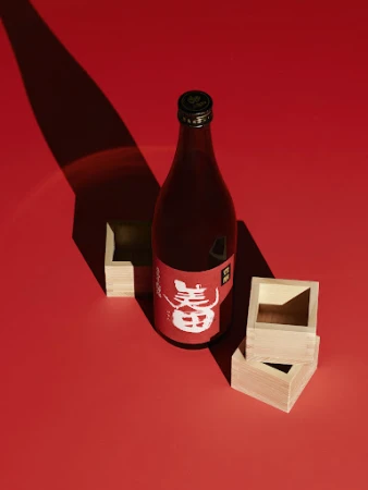 Bottle of sake with wooden square cups on red background. Shadows and light add to the visual interest of the Japanese drink.