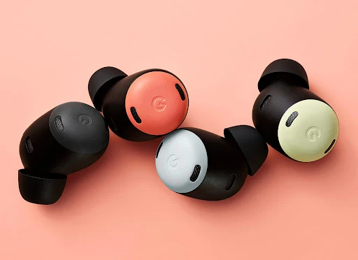 Four black wireless earbuds with a logo rest on a peach-colored surface, displaying multiple colors of the speaker covers.