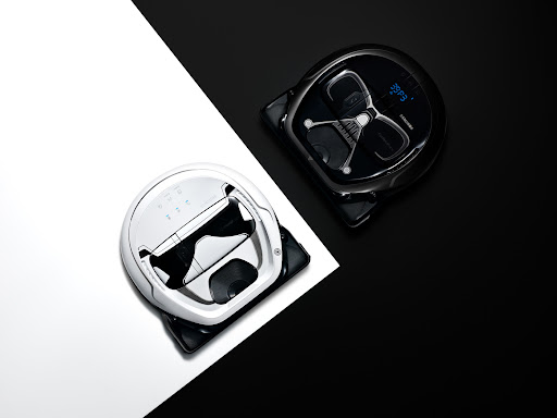 High-angle shot of two robot vacuum cleaners, one white and one black, positioned on a diagonally split black and white background.
