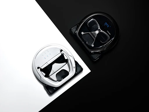 High-angle shot of two robot vacuum cleaners, one white and one black, positioned on a diagonally split black and white background.