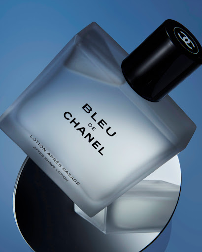 A frosted-glass BLEU de CHANEL bottle with a black cap sits diagonally on a reflective surface against a graduated blue background.