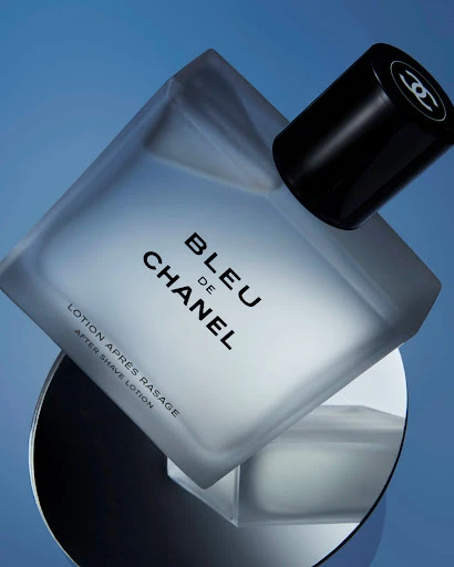 A frosted-glass BLEU de CHANEL bottle with a black cap sits diagonally on a reflective surface against a graduated blue background.