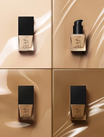 Four bottles of NARS foundation are arranged on a square tile background with beige foundation swirls for a beauty advertisement.