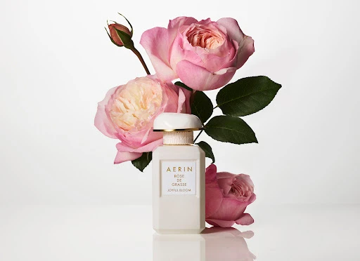 Elegant product shot of Aerin Rose de Grasse perfume bottle surrounded by pink roses against a clean, bright white background.