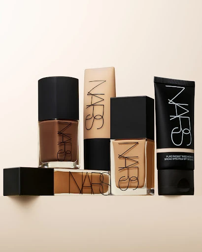 NARS makeup product group on beige background, including foundation in glass bottles and a squeeze tube with black caps and clear logo.