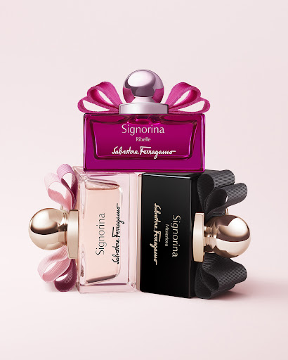 Three 'Signorina' by Salvatore Ferragamo perfume bottles stacked against a light pink backdrop, featuring pink, nude, and black ribbons.