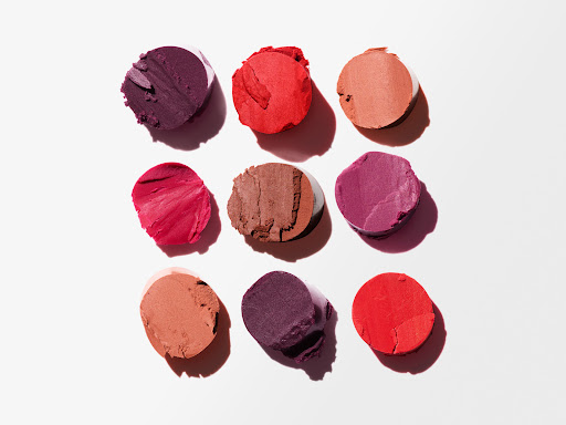 Overhead shot of arranged lipstick smear samples in various red and plum hues on white, suitable for beauty and cosmetics advertising.