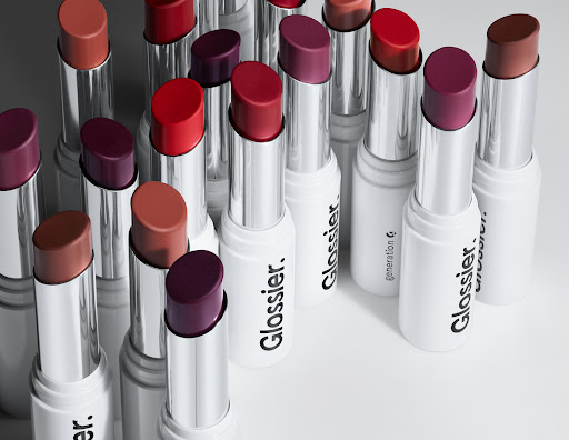 Several rows of multiple shades of Glossier Generation G sheer matte lipstick tubes, showcasing the range of colors available.