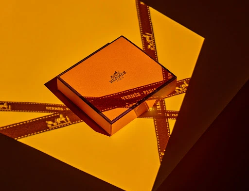 Eye-level shot of a closed orange Hermès box with ribbon on a yellow surface, creating shadows and geometric shapes. 