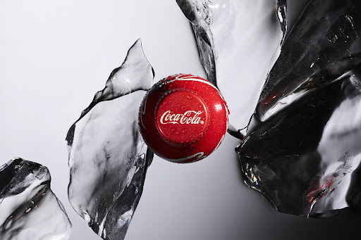 A close-up of a red Coca-Cola drink container surrounded by clear ice shards on a white background, creating a refreshing visual.