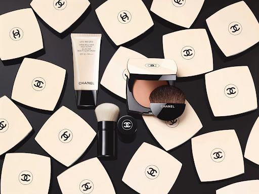 Chanel makeup products arranged on a black surface, featuring powder compacts, a tube of beauty balm, and a makeup brush.