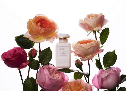 AERIN Rose de Grasse Joyful Bloom perfume bottle surrounded by peach, pink, and red roses with a bright, clean aesthetic.