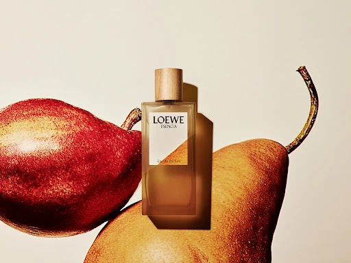 Loewe Esencia perfume bottle with a wood cap, posed with a red pear and yellow pear, on an off-white background, studio lighting.