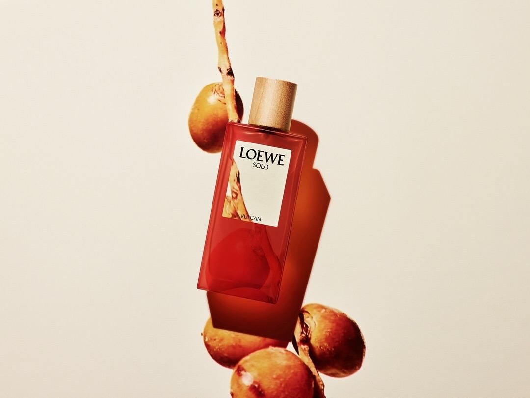 Loewe Solo fragrance bottle with a wooden cap is displayed alongside round, apricot-colored fruits and a thin branch on a light beige background.