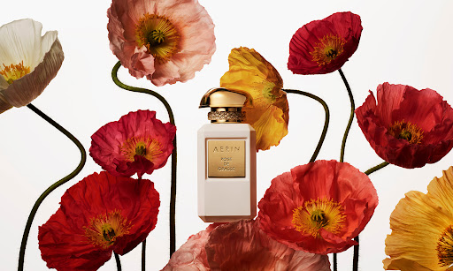 Elegant still life featuring a bottle of Aerin perfume surrounded by vibrant red, yellow, pink, and white poppy flowers on a white background.