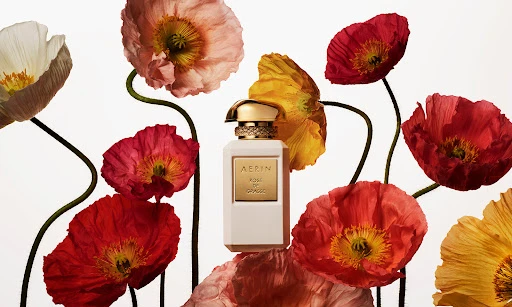 Elegant still life featuring a bottle of Aerin perfume surrounded by vibrant red, yellow, pink, and white poppy flowers on a white background.