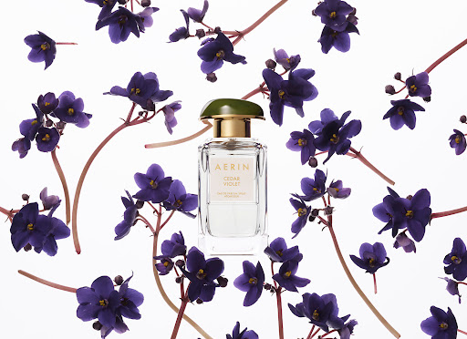 A square AERIN Cedar Violet perfume bottle sits amidst a scattered arrangement of purple violet flowers against a stark white backdrop.