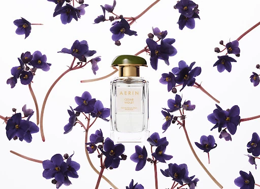 A square AERIN Cedar Violet perfume bottle sits amidst a scattered arrangement of purple violet flowers against a stark white backdrop.