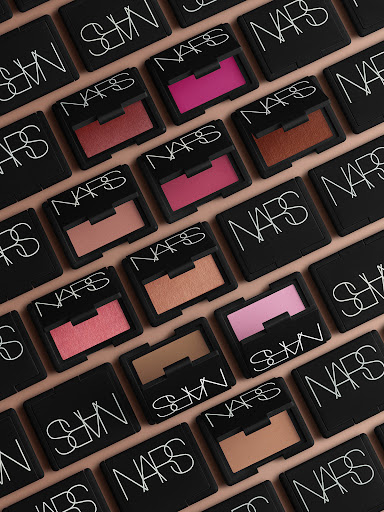 Overhead shot of NARS makeup blush compacts arranged in a grid pattern on a peach-colored background, showcasing various shades of blush.