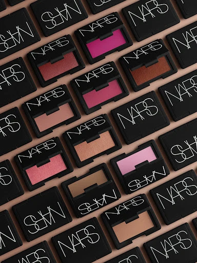 Overhead shot of NARS makeup blush compacts arranged in a grid pattern on a peach-colored background, showcasing various shades of blush.