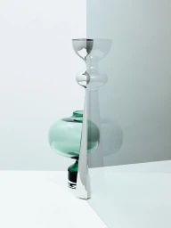 Still life photography of a tall silver candlestick and a green glass vase on a white surface behind a translucent screen.